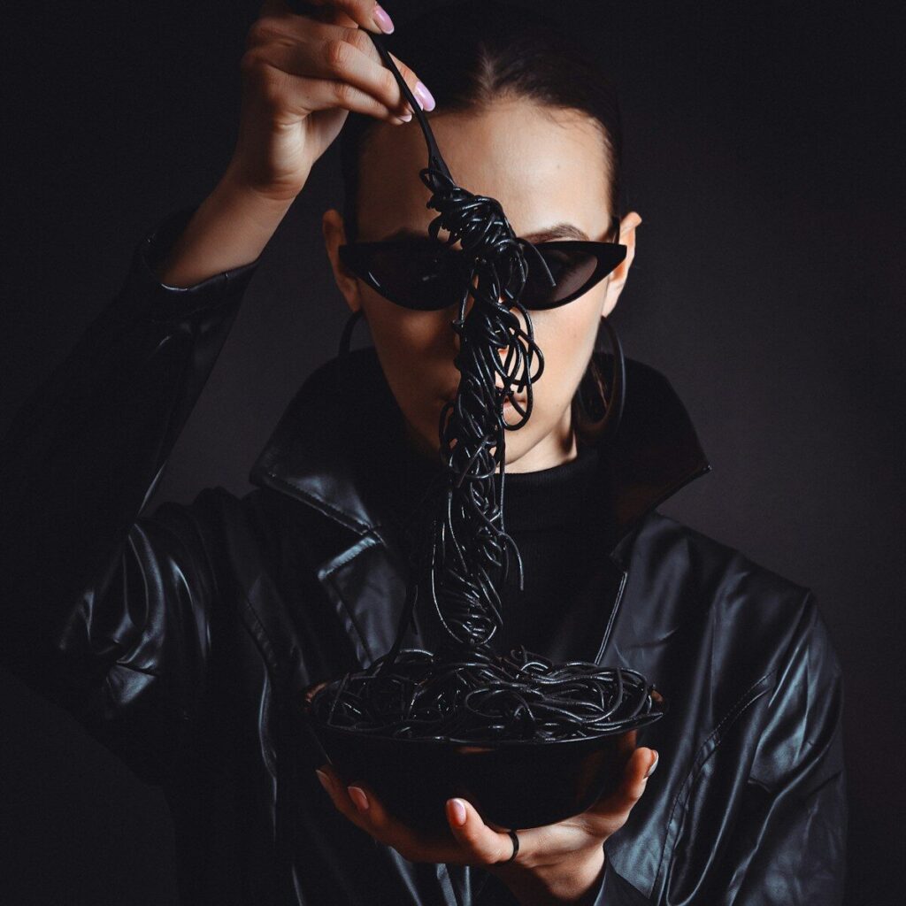 Woman in sunglasses eats black spaghetti from a black bowl.